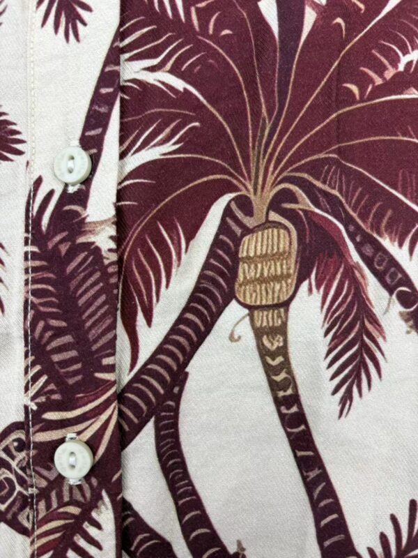 Close-up of a men's shirt with a tropical leaf pattern in shades of burgundy and cream, featuring white buttons.