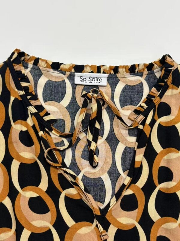 Close-up of a fashionable women's blouse with geometric circle pattern in black, beige, and orange, featuring a tie-neck detail and ruffled collar.