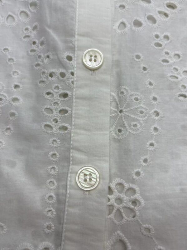 Close-up of a white shirt with intricate eyelet embroidery and two white buttons, showcasing detailed craftsmanship and delicate fabric.