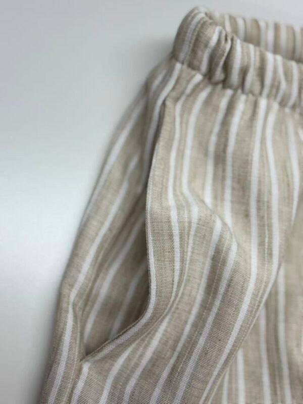 Close-up of beige linen pants with white vertical stripes, elastic waistband, and side pocket, perfect for casual summer wear.