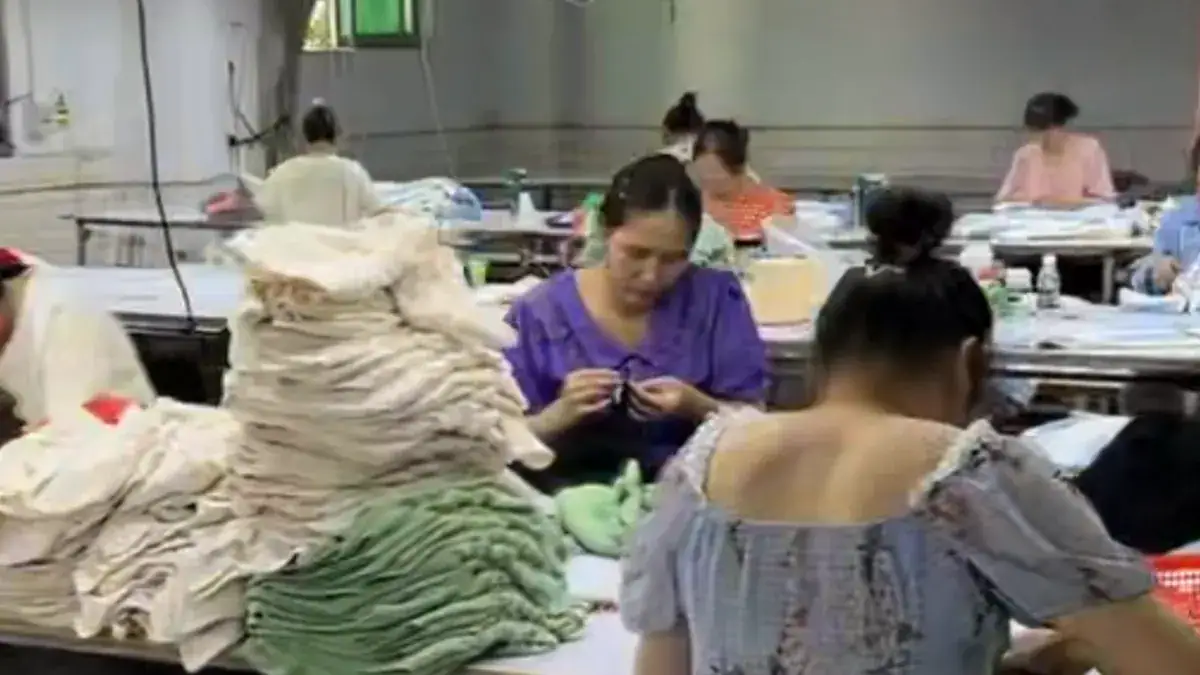 Workers sewing and assembling sweaters in a textile factory, with stacks of finished and unfinished garments around them.