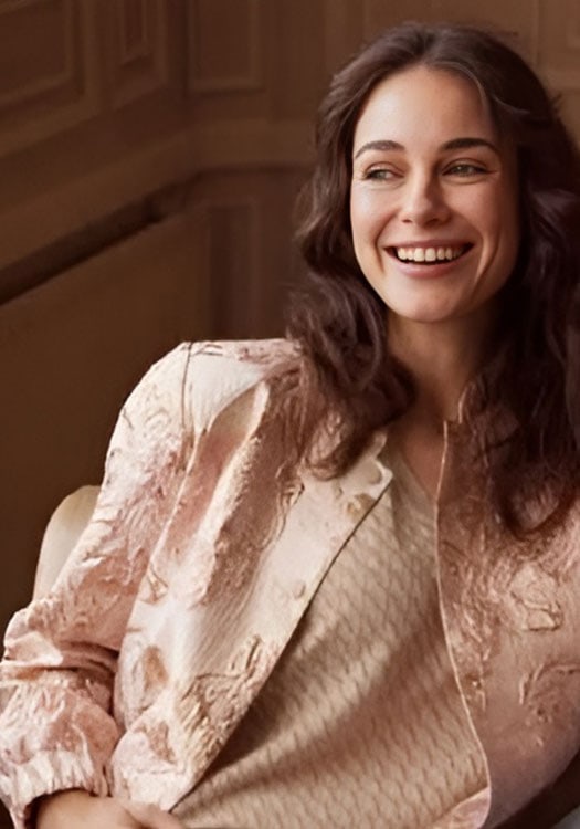 Smiling woman with wavy brown hair wearing a beige blazer and top, sitting in a warmly lit room, showcasing fashion and confidence.