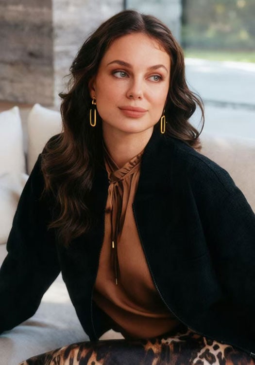Portrait of a woman with wavy brown hair, wearing gold earrings, a brown blouse, and a black jacket, sitting indoors with a blurred background.