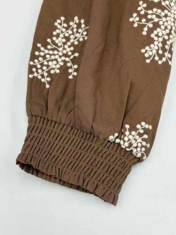 Close-up of brown fabric sleeve with white embroidered floral pattern and elasticated cuff, showcasing detailed craftsmanship.