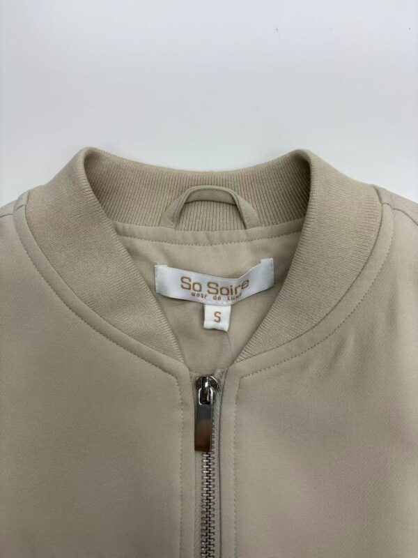 Close-up of a beige zip-up casual jacket for women, featuring a round neckline and a small brand label inside. Perfect for everyday wear and stylish comfort.