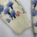 Close-up of a wool sweater with a floral pattern in blue, black, and pink on a cream background, showcasing detailed knitting and vibrant colors.