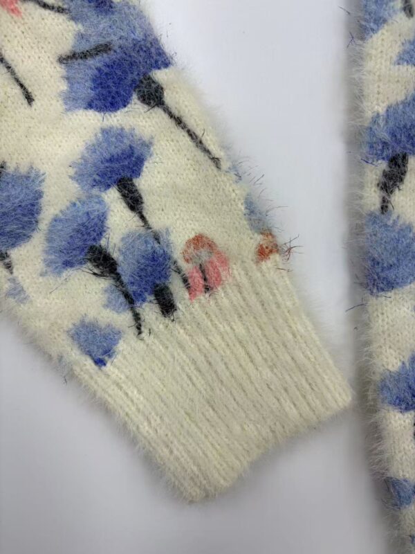 Close-up of a wool sweater with a floral pattern in blue, black, and pink on a cream background, showcasing detailed knitting and vibrant colors.