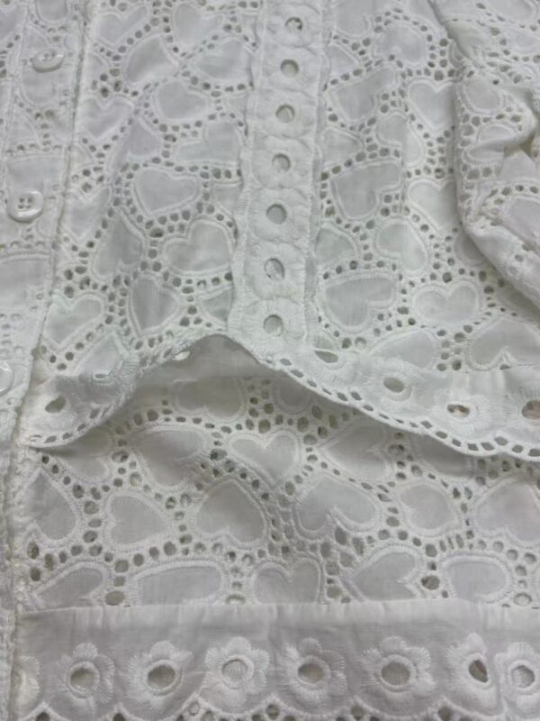 Close-up of white eyelet lace fabric with intricate heart and floral patterns, showcasing detailed embroidery and cutwork for fashion and textile design.