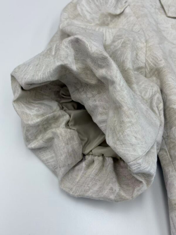 Close-up of a light beige fabric garment with intricate texture and subtle pattern, showcasing high-quality material and craftsmanship.