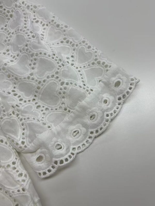 Close-up of white eyelet lace fabric with intricate heart-shaped cutouts and embroidered details, perfect for fashion and home decor.