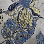 Close-up of intricate embroidered floral pattern with blue, gold, and silver threads on a light fabric background.
