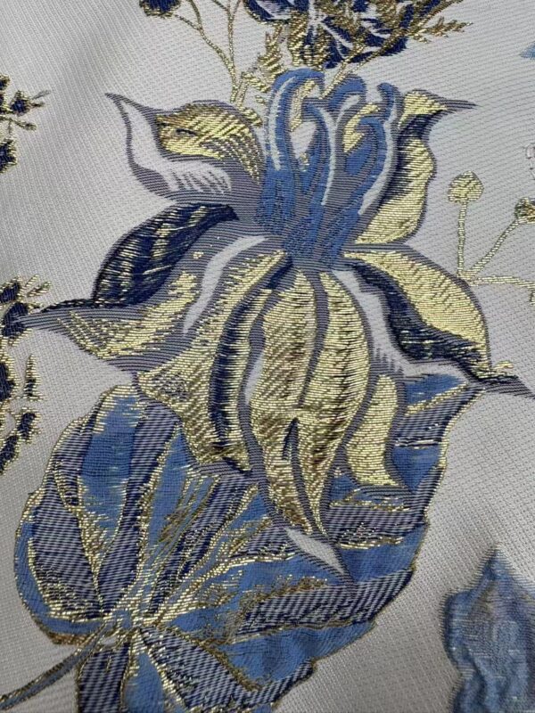 Close-up of intricate embroidered floral pattern with blue, gold, and silver threads on a light fabric background.