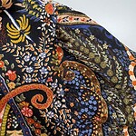 Close-up of vibrant embroidered fabric with floral and paisley patterns in blue, orange, yellow, and green on black background.