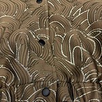 Close-up of a brown fabric with intricate silver and gold swirling patterns, featuring black buttons and a ruffled hem, suitable for stylish clothing.