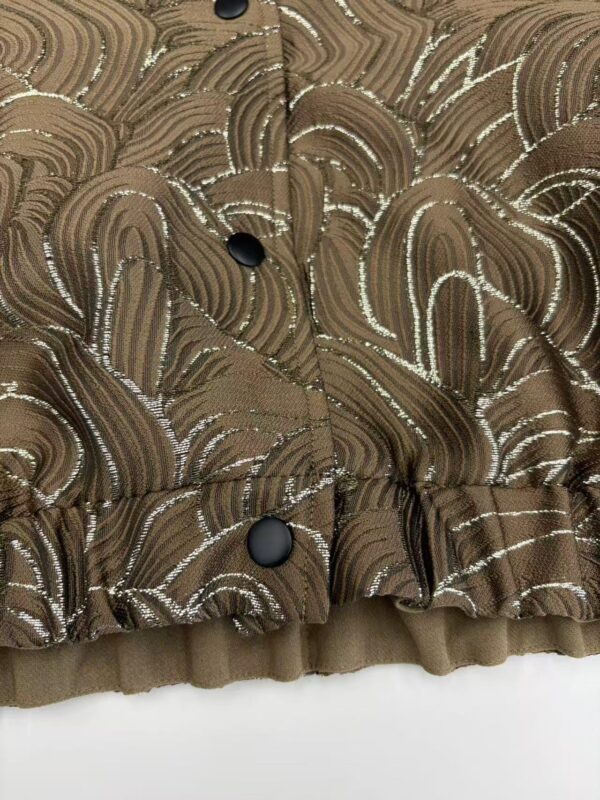 Close-up of a brown fabric with intricate silver and gold swirling patterns, featuring black buttons and a ruffled hem, suitable for stylish clothing.