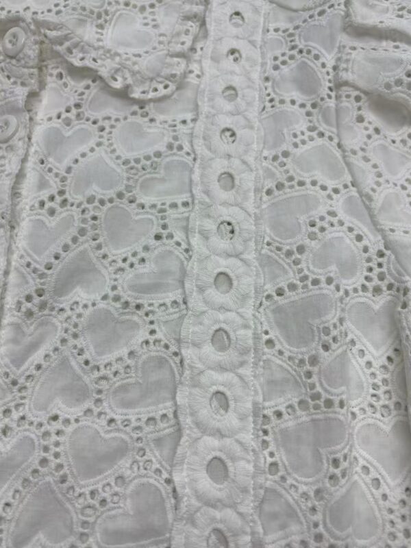 Close-up of white eyelet lace fabric with heart-shaped cutouts and embroidered details, showcasing intricate craftsmanship and delicate design.