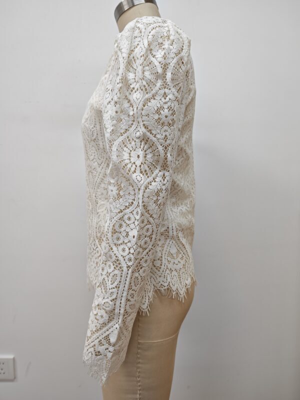 Close-up of a white lace dress with intricate floral patterns, long sleeves, and scalloped edges, displayed on a mannequin against a plain background.