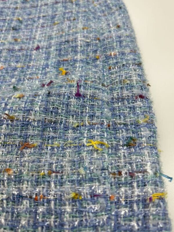 Close-up of a textured tweed fabric featuring a mix of blue, white, and multicolored threads, showcasing detailed weave and colorful accents.