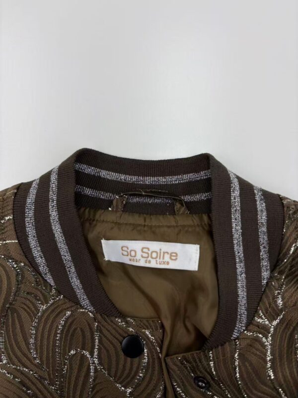Close-up of a brown bomber jacket with intricate embroidery and striped ribbed collar, featuring a label that reads 'So Soire' for fashion and style.