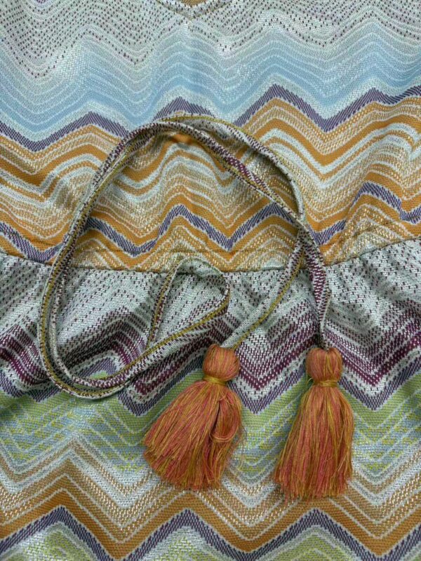 Vibrant handwoven textile featuring zigzag patterns in blue, yellow, purple, and green with decorative tassels in orange and pink.